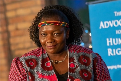[ai] A woman with curly hair wearing a colorful headband and a patterned jacket with circular designs, seated in front of a blue banner that reads "Advocacy for Human Rights".